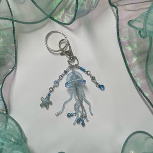 Puede incluir: Un llavero de medusa de vidrio azul claro con cuentas plateadas y un dije de estrella de mar plateada. El llavero est&aacute; colgado de un anillo de llave plateado.