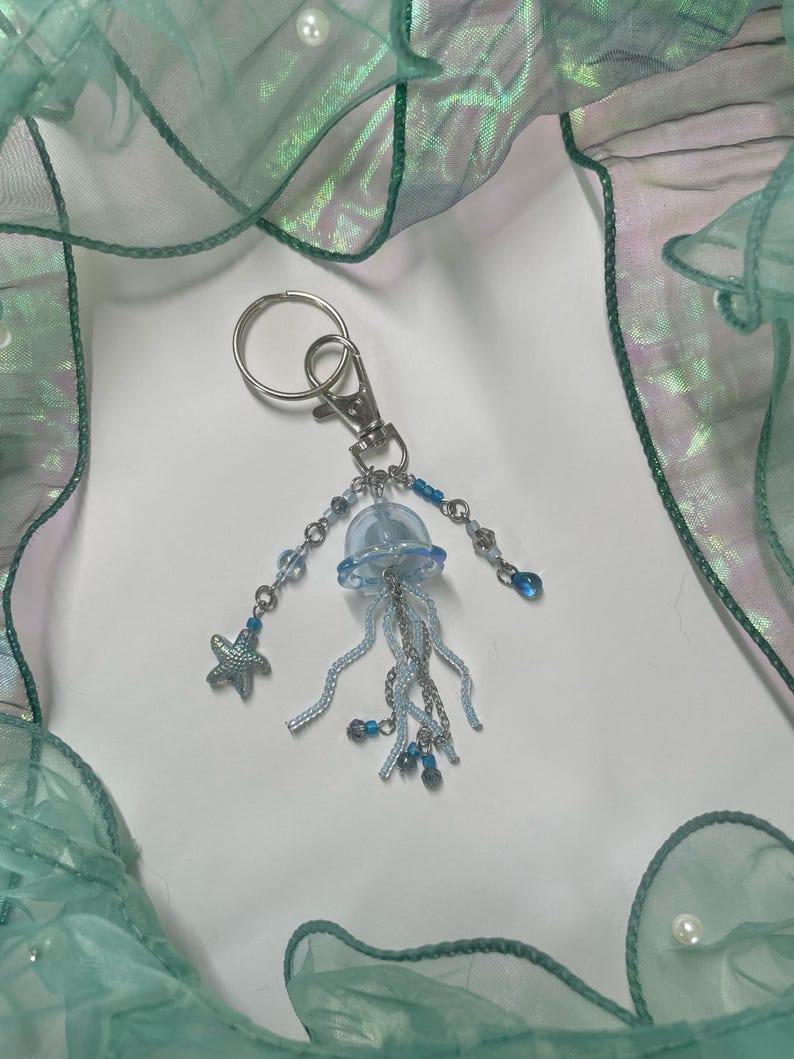 Puede incluir: Un llavero de medusa de vidrio azul claro con cuentas plateadas y un dije de estrella de mar plateada. El llavero est&aacute; colgado de un anillo de llave plateado.