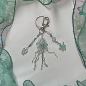 Puede incluir: Un llavero de plata con un dije de medusa transparente e iridiscente, un dije de pez plateado, un dije de estrella plateada y cuentas plateadas.