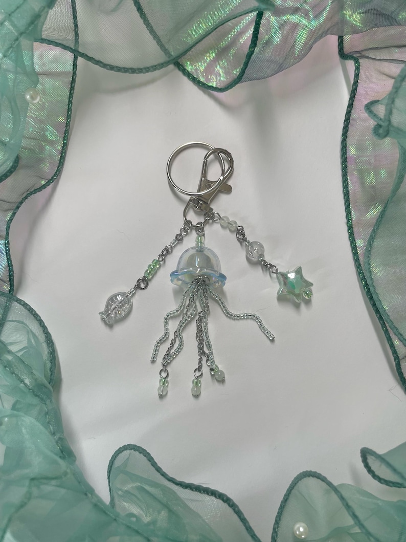 Puede incluir: Un llavero de plata con un dije de medusa transparente e iridiscente, un dije de pez plateado, un dije de estrella plateada y cuentas plateadas.