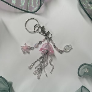 Puede incluir: Un charm de medusa rosa con cadenas y cuentas plateadas, unido a un llavero plateado. El charm incluye una estrella de mar rosa y una cuenta de vidrio transparente.