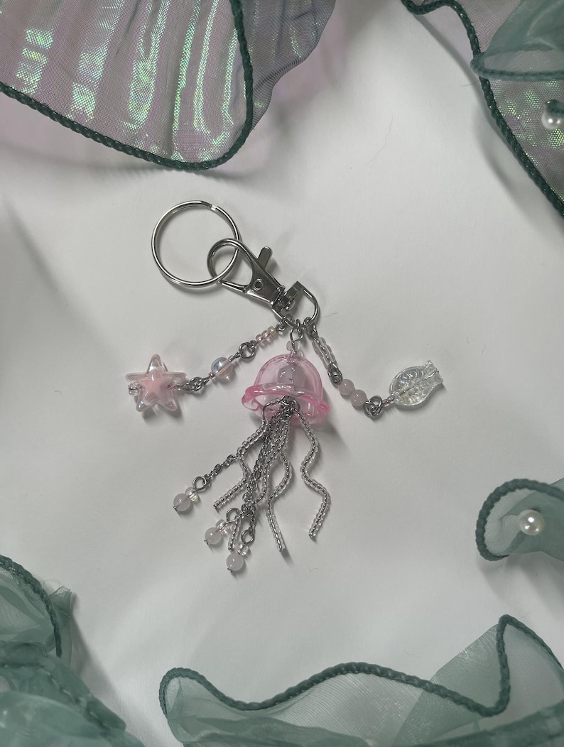 Puede incluir: Un charm de medusa rosa con cadenas y cuentas plateadas, unido a un llavero plateado. El charm incluye una estrella de mar rosa y una cuenta de vidrio transparente.