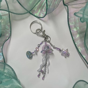 Puede incluir: Un llavero plateado con un dije de medusa iridiscente morado, un dije de estrella plateada y un dije de concha plateada. El llavero est&aacute; colgado de un anillo plateado.