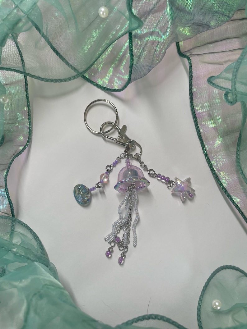 Puede incluir: Un llavero plateado con un dije de medusa iridiscente morado, un dije de estrella plateada y un dije de concha plateada. El llavero est&aacute; colgado de un anillo plateado.