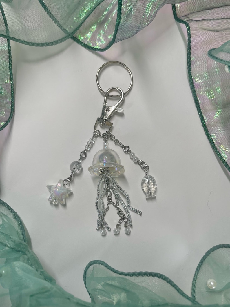 Puede incluir: Un llavero plateado con un dije de medusa transparente iridiscente, un dije de estrella plateada y un dije de concha plateada. Los dijes est&aacute;n unidos a una cadena plateada con peque&ntilde;as cuentas blancas.