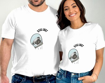 May include: White t-shirts with a cartoon pug wearing a space helmet. The text "STILL LOST" is above the pug illustration. The shirts are worn by two people, one male and one female, both wearing blue jeans.