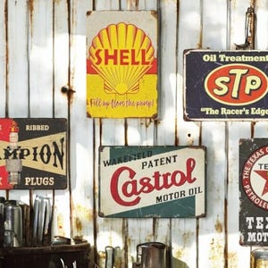 May include: Five vintage metal signs with a rusty, weathered look. The signs advertise automotive products like Shell, Champion Spark Plugs, Castrol Motor Oil, STP Oil Treatment, and Texaco Motor Oil. The signs are displayed on a white, corrugated metal wall.