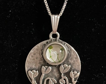 May include: A silver necklace with a round pendant featuring a pressed flower design. The pendant has a textured surface and a clear glass dome over the flower.