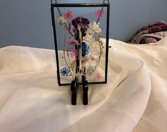 Könnte beinhalten: Ein gerahmter gepresster Blumenstrauß mit einer Auswahl getrockneter Blumen in den Farben Rosa, Lila, Blau und Weiß. Die Blumen sind in einem Glasrahmen mit schwarzem Metallrahmen und Ständer angeordnet.