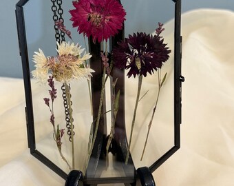 Könnte beinhalten: Ein achteckiger Glasrahmen mit einer schwarzen Metallkette, die oben hängt. Im Rahmen befinden sich drei gepresste Blumen: eine weiße Blume, eine dunkelviolette Blume und eine rosa Blume. Der Rahmen steht auf einem schwarzen Staffelei.