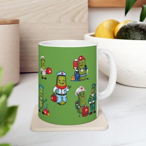May include: White ceramic mug featuring a green background with cartoon pickle characters dressed as medical professionals. Each pickle is wearing a different uniform and carrying a red first aid kit. The mug has a white handle and is sitting on a beige coaster.