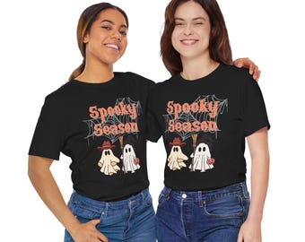 May include: Two people wearing black t-shirts with the words "Spooky Season" in orange script above a graphic of two ghosts and a witch. The ghosts are holding a pumpkin and a broom. The t-shirts are paired with blue jeans.