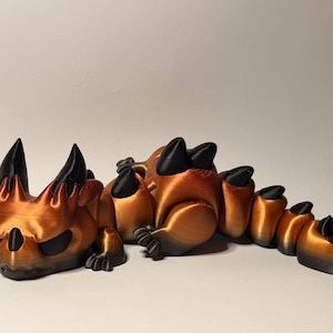 May include: A 3D-printed articulated dragon figurine in shades of bronze and black. The dragon has a detailed, stylized design with sharp horns and claws. The figurine is posed in a curved, serpentine position, showcasing its segmented body.
