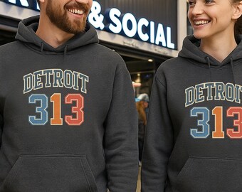 Peut inclure: Deux personnes portent des sweats à capuche gris foncé avec le mot "DETROIT" et le numéro "313" dans une police rétro. Les sweats à capuche ont une poche avant et une capuche à cordon. En arrière-plan, une enseigne indique "SPORTS & SOCIAL".