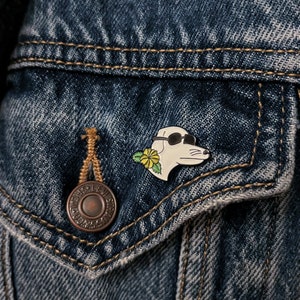 May include: A white enamel pin of a dog wearing sunglasses and a yellow flower. The pin is attached to a denim jacket.