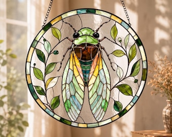 May include: A circular stained glass suncatcher featuring a central cicada with iridescent green, yellow, and brown wings. The insect is surrounded by delicate green glass leaves and vines, all encased in a dark metal frame with a hanging chain.