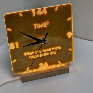 May include: A yellow acrylic clock with black hands and a white inscription that reads "144 TIME2 When you need more hours in the day". The clock is on a wooden base.
