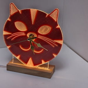 May include: A red cat-shaped clock with gold hands and a wooden base. The clock is illuminated from behind, creating a warm glow. The base is inscribed with "Dows VC-2023".
