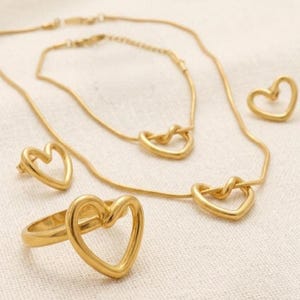 May include: A gold-toned jewelry set consisting of a necklace with a heart-shaped pendant, a matching bracelet with a heart-shaped charm, a pair of heart-shaped stud earrings, and a ring featuring a prominent open heart design on the band.