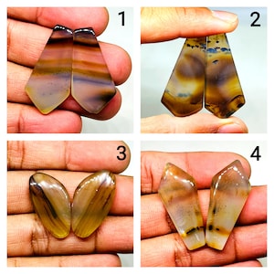 May include: Four pairs of polished gemstone pendants in various shapes. Each pair features a unique pattern of brown, tan, and cream colours. The gemstones are held in hands, showcasing their natural beauty and variations.
