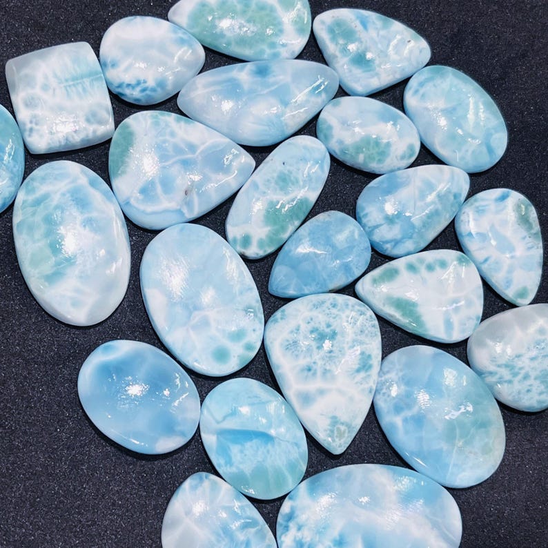 May include: A collection of polished larimar gemstones in various shapes, including oval, teardrop, and rectangular. The stones display a range of blue and white hues, with patterns resembling ocean waves. The gemstones are arranged on a dark surface.