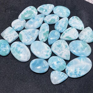 May include: A collection of polished larimar gemstones in various shapes and sizes. The stones display a range of blue and white hues, with some featuring green accents. The gemstones are smooth and have a natural, organic appearance.
