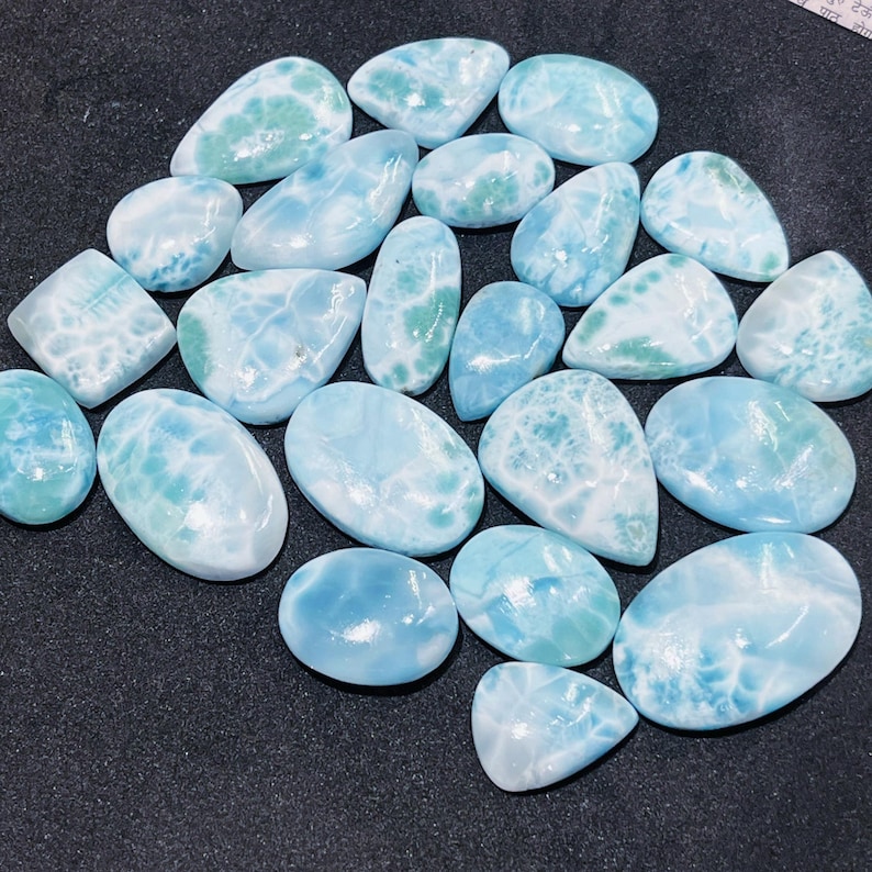 May include: A collection of polished larimar gemstones in various shapes and sizes. The stones display a range of blue and white hues, with some featuring green accents. The gemstones are smooth and have a natural, organic appearance.