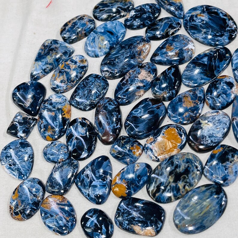 May include: A selection of polished gemstones in diverse shapes, such as oval and teardrop. The stones display a deep blue base colour with swirling patterns of white and golden-brown inclusions, creating a marbled effect. The gemstones are presented on a white surface.