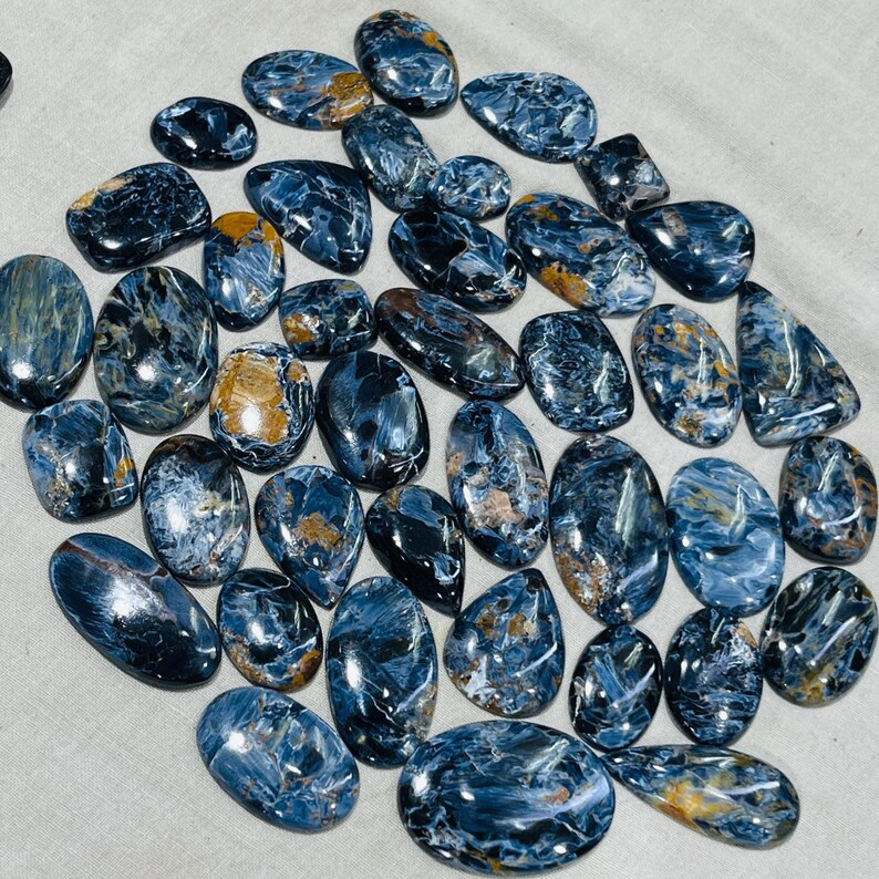 May include: A selection of polished pietersite gemstones in diverse shapes, such as oval, teardrop, and irregular. The stones exhibit a blend of deep blue, grey, and golden-brown hues, with a glossy finish. The gemstones are arranged on a light-coloured surface.