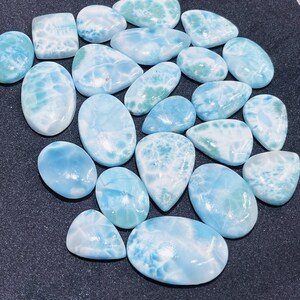 May include: A collection of polished larimar gemstones in various shapes, including oval, teardrop, and rectangular. The stones display a range of light blue and white patterns, with some showing hints of green. The gemstones are arranged on a dark surface.