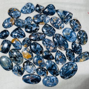 May include: A selection of polished pietersite gemstones in diverse shapes. The stones exhibit a blend of deep blue, grey, and golden-brown tones, featuring swirling patterns and a glossy finish. The gemstones are displayed on a light-coloured surface.
