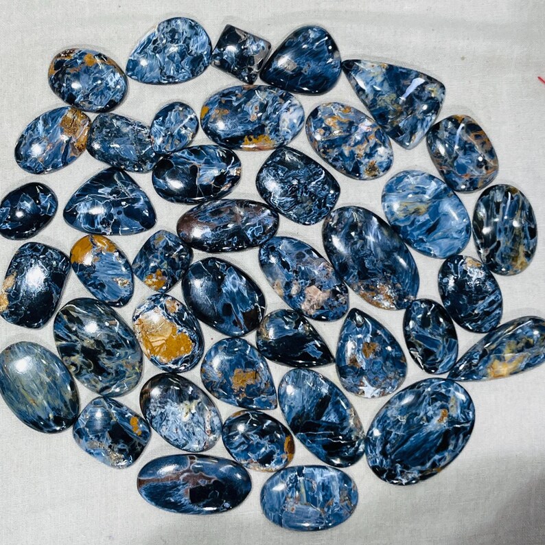May include: A selection of polished pietersite gemstones in diverse shapes. The stones exhibit a blend of deep blue, grey, and golden-brown tones, featuring swirling patterns and a glossy finish. The gemstones are displayed on a light-coloured surface.