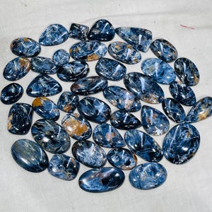 May include: A selection of polished Pietersite gemstones in diverse shapes, such as oval and teardrop. The stones exhibit a blend of deep blue, grey, and golden-brown hues, with swirling patterns and a glossy finish. The gemstones are arranged on a light-coloured surface.