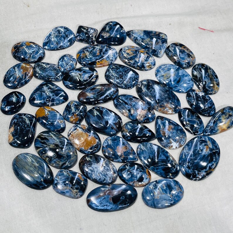 May include: A selection of polished Pietersite gemstones in diverse shapes, such as oval and teardrop. The stones exhibit a blend of deep blue, grey, and golden-brown hues, with swirling patterns and a glossy finish. The gemstones are arranged on a light-coloured surface.