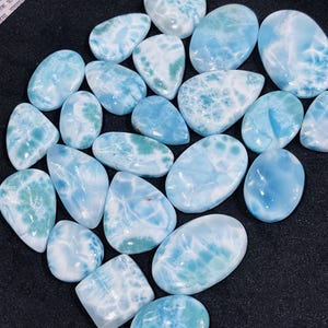May include: A collection of polished larimar gemstones. The stones vary in shape, including oval and teardrop, and feature a mix of light blue and white patterns. Some stones have hints of green.