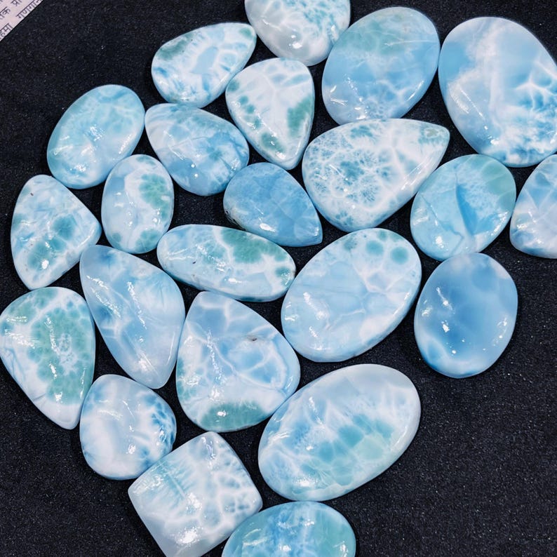 May include: A collection of polished larimar gemstones. The stones vary in shape, including oval and teardrop, and feature a mix of light blue and white patterns. Some stones have hints of green.