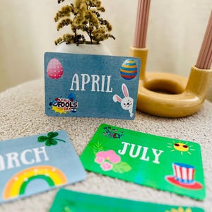 May include: Blue, green, and white cards with the months of April, July, and June written on them. Each card has a different design and illustration. April has an Easter egg and bunny, July has a Fourth of July theme, and June has a rainbow.