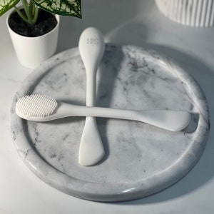 May include: Two white silicone facial cleansing tools on a white marble surface. One tool has a brush on one end and a flat edge on the other. The other tool has a flat edge on both ends.