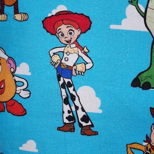 May include: A blue fabric featuring a repeating pattern of Toy Story characters including Jessie the cowgirl with her red hat, white shirt, and cow-print pants, alongside parts of other characters like a green dinosaur and a potato figure.