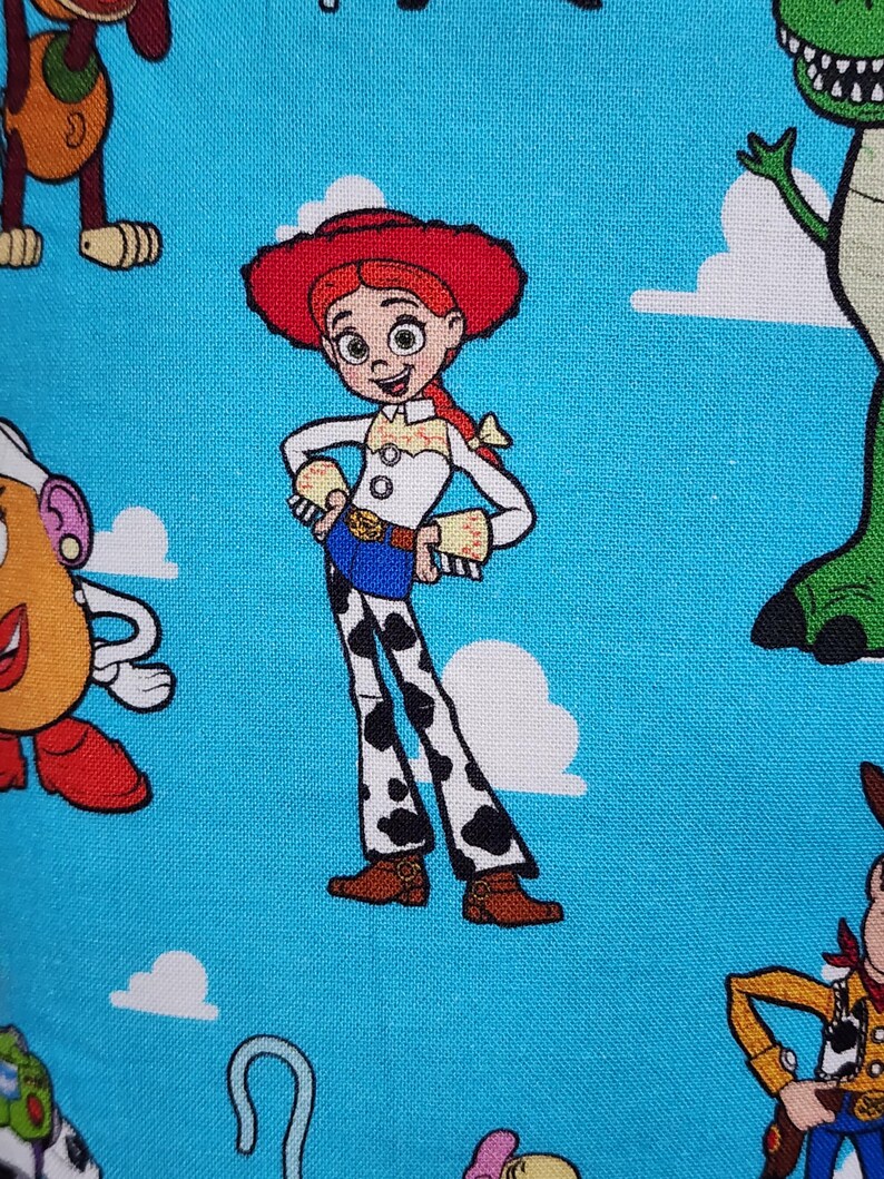 May include: A blue fabric featuring a repeating pattern of Toy Story characters including Jessie the cowgirl with her red hat, white shirt, and cow-print pants, alongside parts of other characters like a green dinosaur and a potato figure.