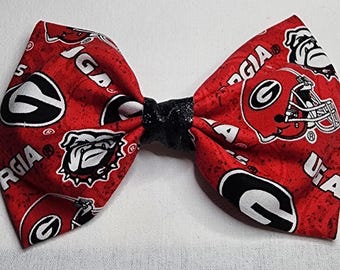 May include: A red and black bow tie featuring the University of Georgia Bulldogs logo and helmet. The bow tie has a black center and is made of fabric. The bow tie is a fan accessory.