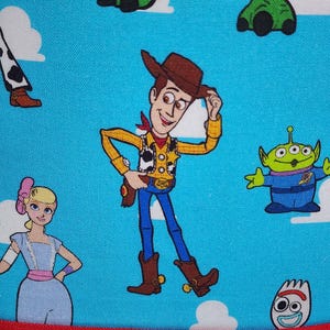 May include: A bright blue fabric featuring a repeating pattern of Toy Story characters including Woody the cowboy, Bo Peep, a green alien, and Forky, set against a background of white clouds. The bottom edge is finished with a solid red fabric border.