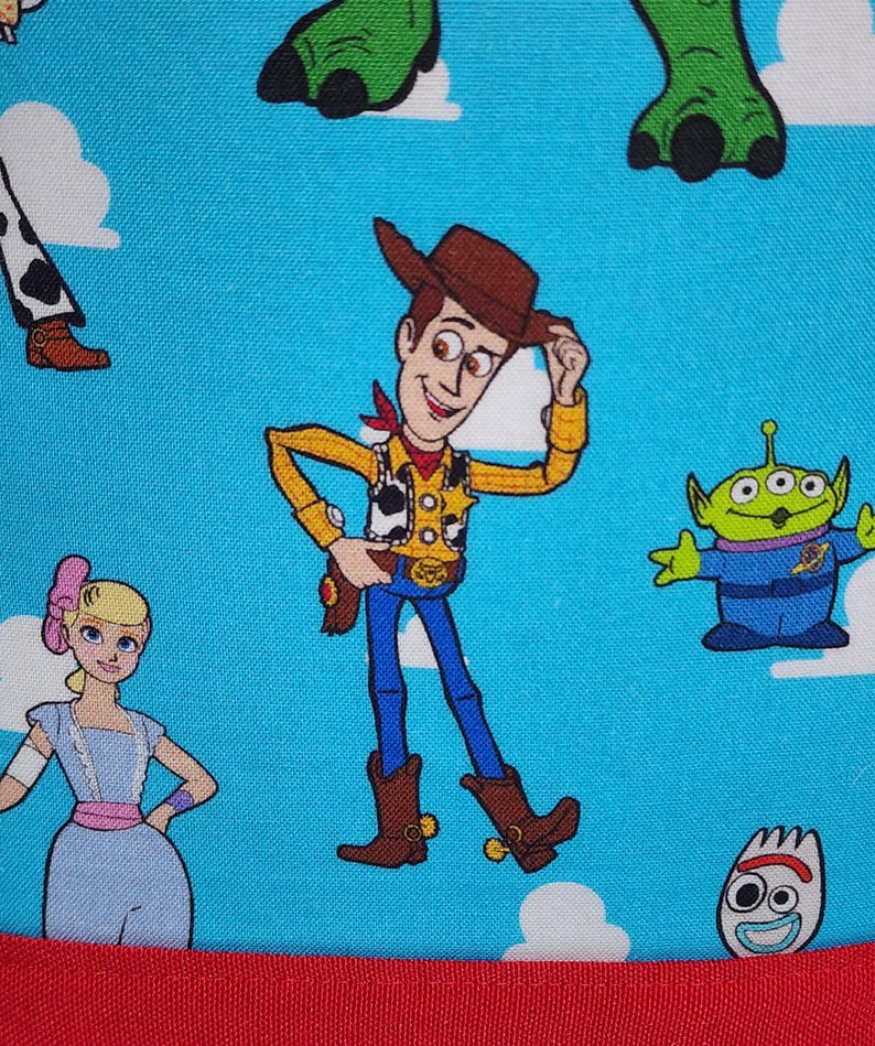 May include: A bright blue fabric featuring a repeating pattern of Toy Story characters including Woody the cowboy, Bo Peep, a green alien, and Forky, set against a background of white clouds. The bottom edge is finished with a solid red fabric border.