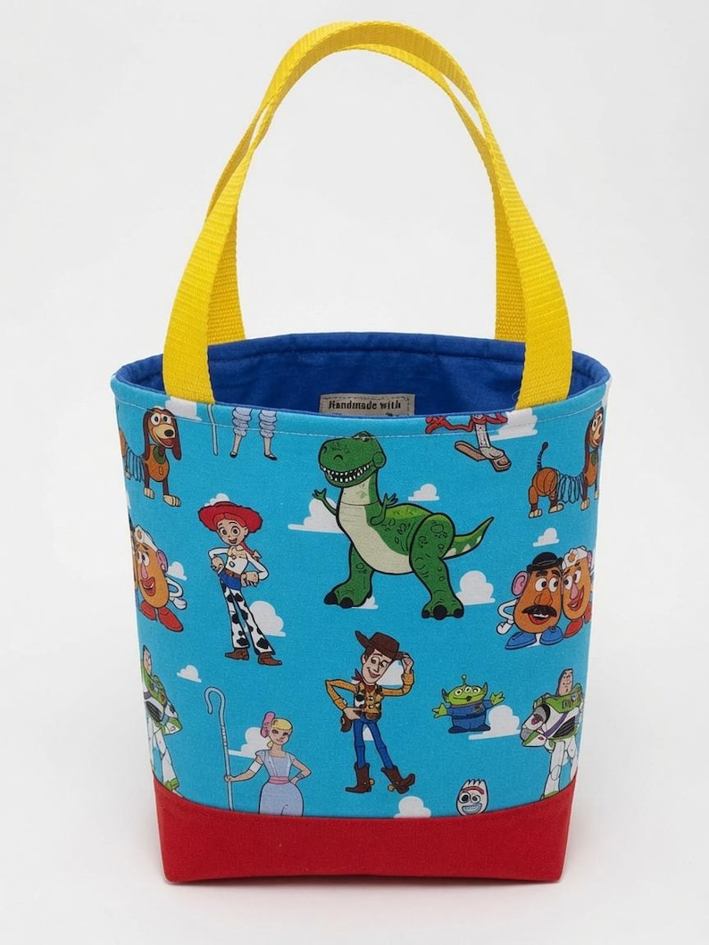 May include: A blue fabric tote bag featuring a repeating pattern of Toy Story characters including Woody, Buzz Lightyear, Jessie, Rex, and Slinky Dog against a sky blue background with white clouds. The bag has yellow handles and a solid red base.