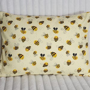 May include: A rectangular pillow with a cream-colored background, covered in a repeating pattern of cartoon bees. The bees are yellow and black with smiling faces and small wings. The pillow is a standard size.