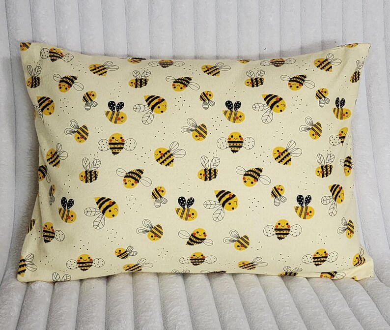 May include: A rectangular pillow with a cream-colored background, covered in a repeating pattern of cartoon bees. The bees are yellow and black with smiling faces and small wings. The pillow is a standard size.