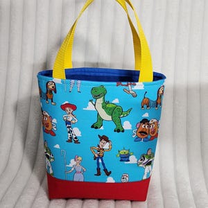 May include: A fabric tote bag featuring a light blue background with a repeating pattern of Toy Story characters including Woody, Jessie, Rex, and Buzz Lightyear. The bag has a solid red base, a dark blue interior lining, and two bright yellow nylon handles.