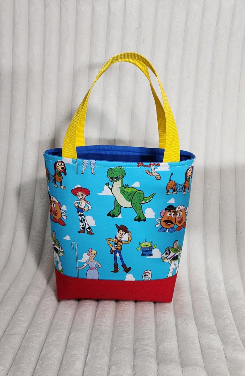 May include: A fabric tote bag featuring a light blue background with a repeating pattern of Toy Story characters including Woody, Jessie, Rex, and Buzz Lightyear. The bag has a solid red base, a dark blue interior lining, and two bright yellow nylon handles.