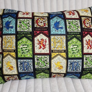 May include: Rectangular pillow with a colorful stained-glass design featuring the crests of Hogwarts houses: Gryffindor, Slytherin, Hufflepuff, and Ravenclaw. The pillow has a black background and is filled with various colors.