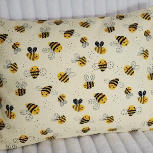 May include: Rectangular pillow with a cream-colored background covered in a pattern of yellow and black cartoon bees. The bees have smiling faces and are surrounded by small black dots.
